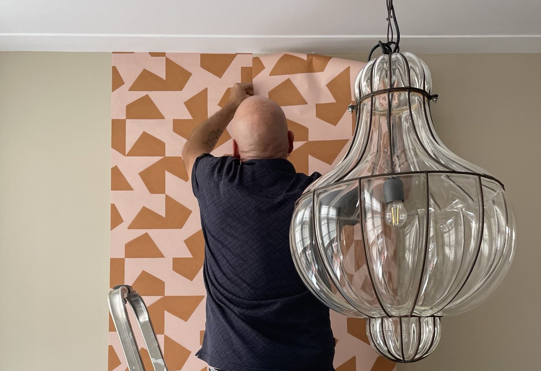 Leen Fischer brengt behang met een oranje grafisch patroon aan naast een glazen hanglamp